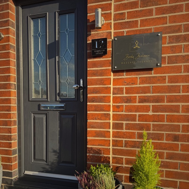 A brick exterior entrance with a dark front door featuring decorative glass panels. Next to the door, a mounted sign reads “Body Balance Massage Therapy,” indicating the location of the practice. A small wall light fixture is installed above, and potted plants at the base add a welcoming, well-kept appearance.